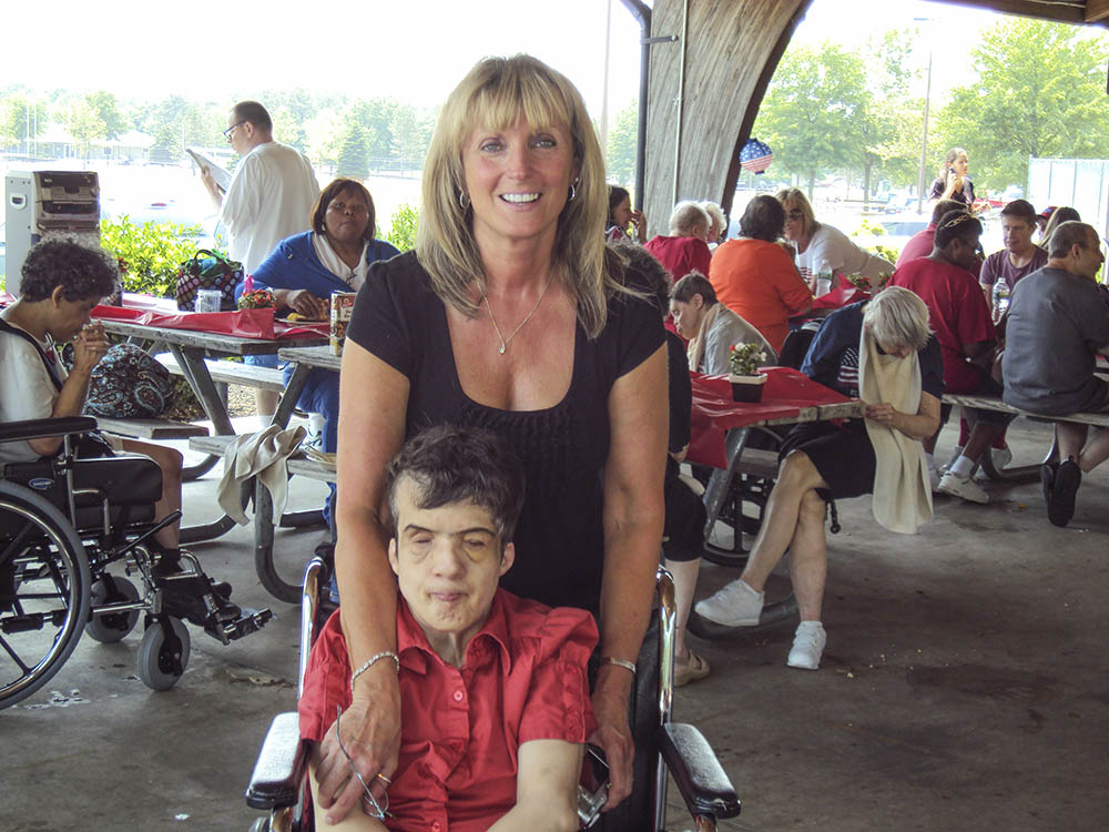 A staff member of AMIB stands behind a resident in a wheel chair.